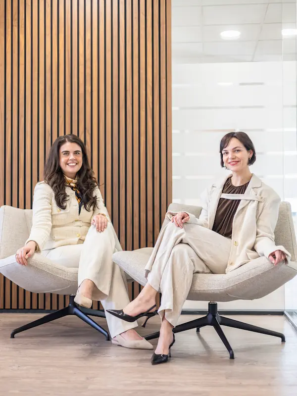 Twee huidtherapeuten van HuidSkills Kliniek zitten ontspannen in moderne fauteuils in een stijlvolle kliniekruimte met houten wandpanelen en glazen wanden op de achtergrond.