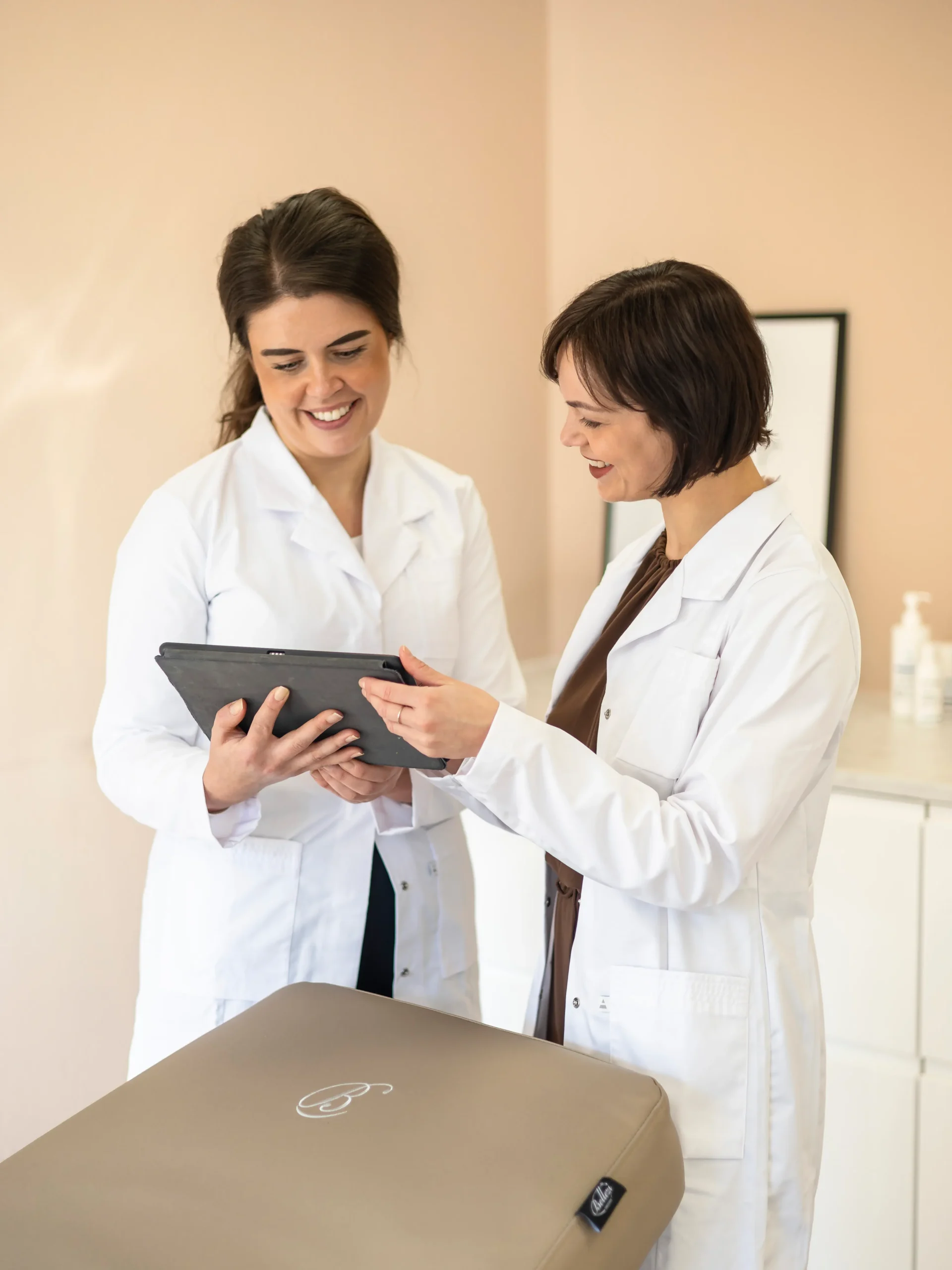 Twee huidtherapeuten in witte jassen bekijken samen een tablet in een behandelruimte