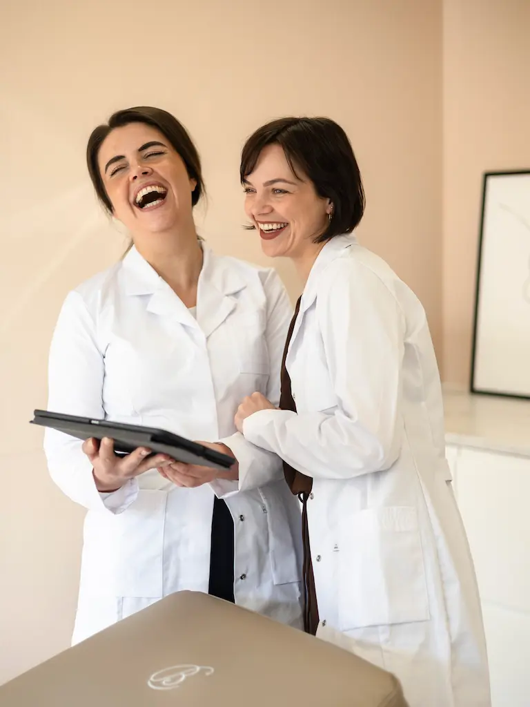 Twee huidtherapeuten van HuidSkills Kliniek in witte jassen lachen samen terwijl ze een tablet bekijken in een lichte behandelruimte voor huidverbetering.