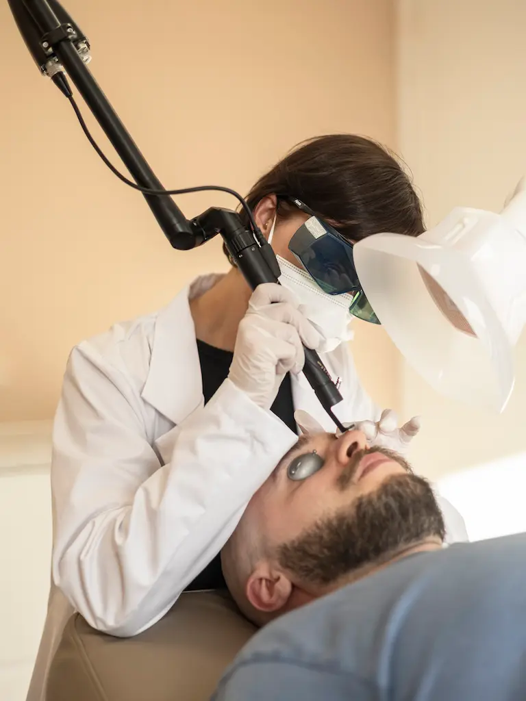 Huidtherapeut voert een gerichte laserbehandeling uit op het gezicht van een mannelijke cliënt die beschermende oogpads draagt tijdens de behandeling.